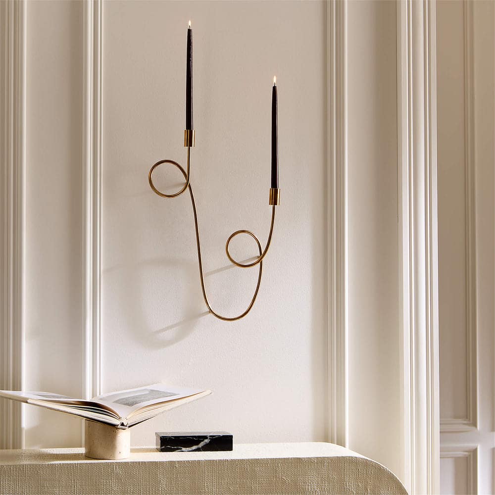 A sculptural brass candle holder wall sconce with two curved arms holds tall, thin black candles. Below, an open book rests on a textured beige surface against a white paneled wall.