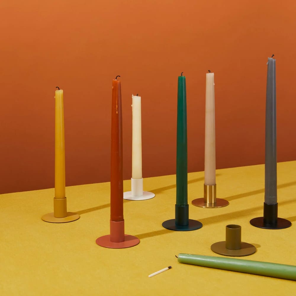 Seven colorful taper candles, each in a different candle holder, are arranged on a yellow surface with an orange background. In the foreground, a green candle and matching candle holder rest beside a matchstick on the table.
