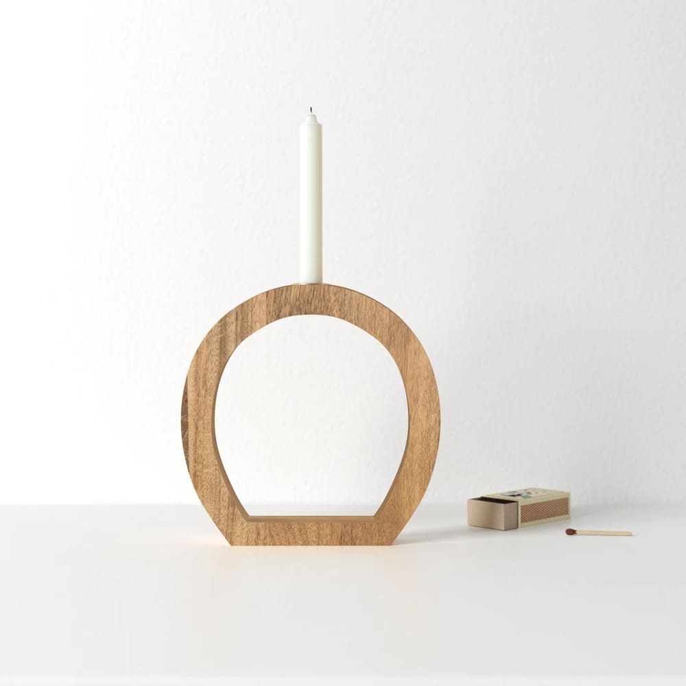 A minimalist candle holder, crafted from wood and shaped like an open circle, holds a white taper candle. A matchbox and a single match rest nearby on the white surface, all set against a plain white background.