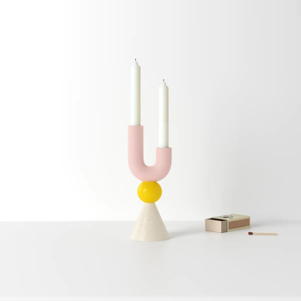 A modern candle holder with a pink U-shaped design, yellow sphere, and white cone base holds two white candles. The candle holder rests on a plain white surface beside a matchbox and match, set against a white background.