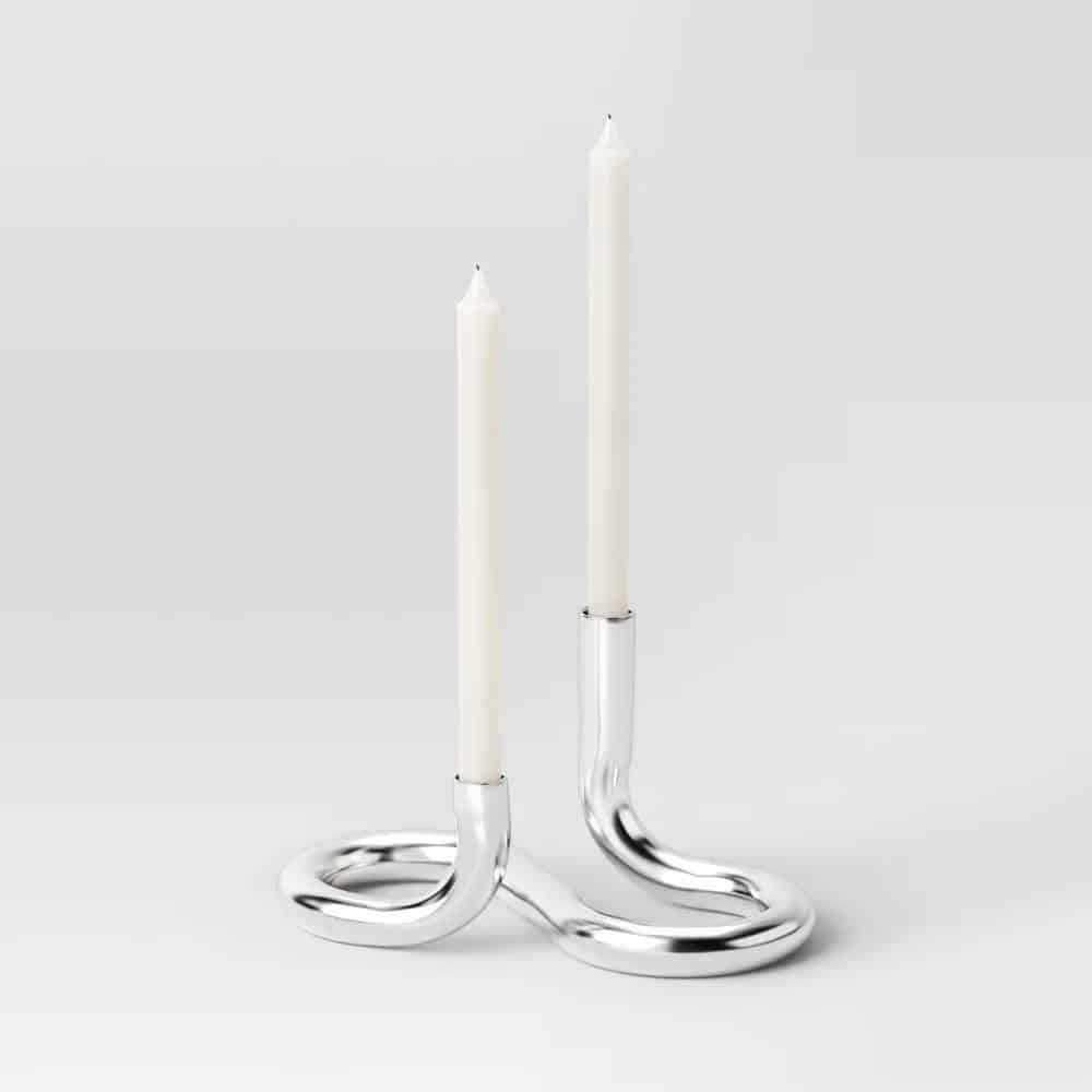 A modern silver candle holder with two curved, looping arms, each elegantly holding a tall white taper candle against a plain light gray background.