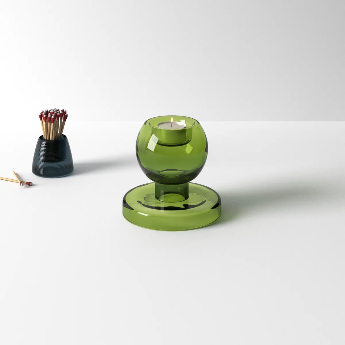 A green glass candle holder with a tealight rests on a white surface, accompanied by a small dark container holding red-tipped matches nearby.