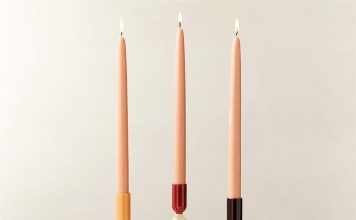 Three lit peach-colored taper candles in uniquely shaped candle holders—brown with wavy design, clear with slender base, and red with geometric design—are arranged side by side on a white surface.