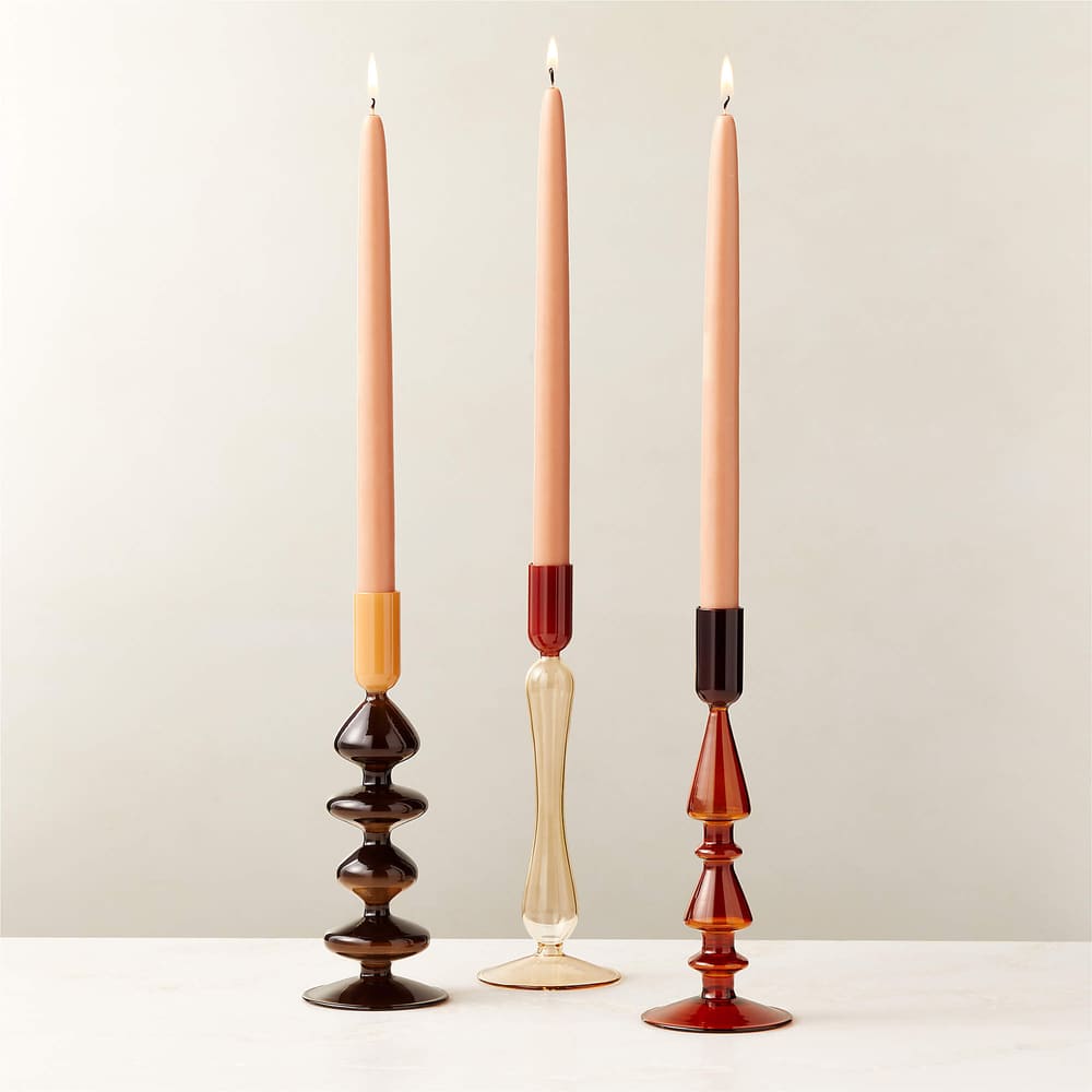 Three lit peach-colored taper candles in uniquely shaped candle holders—brown with wavy design, clear with slender base, and red with geometric design—are arranged side by side on a white surface.