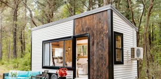 A stylish tiny house on wheels with white siding, dark wood accents, and large windows sits in a forest. A small deck leads to the door, with outdoor seating featuring turquoise cushions and a table nearby.