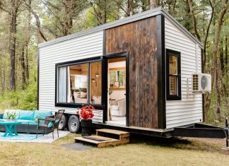A stylish tiny house on wheels with white siding, dark wood accents, and large windows sits in a forest. A small deck leads to the door, with outdoor seating featuring turquoise cushions and a table nearby.