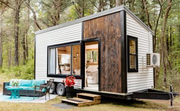 A stylish tiny house on wheels with white siding, dark wood accents, and large windows sits in a forest. A small deck leads to the door, with outdoor seating featuring turquoise cushions and a table nearby.