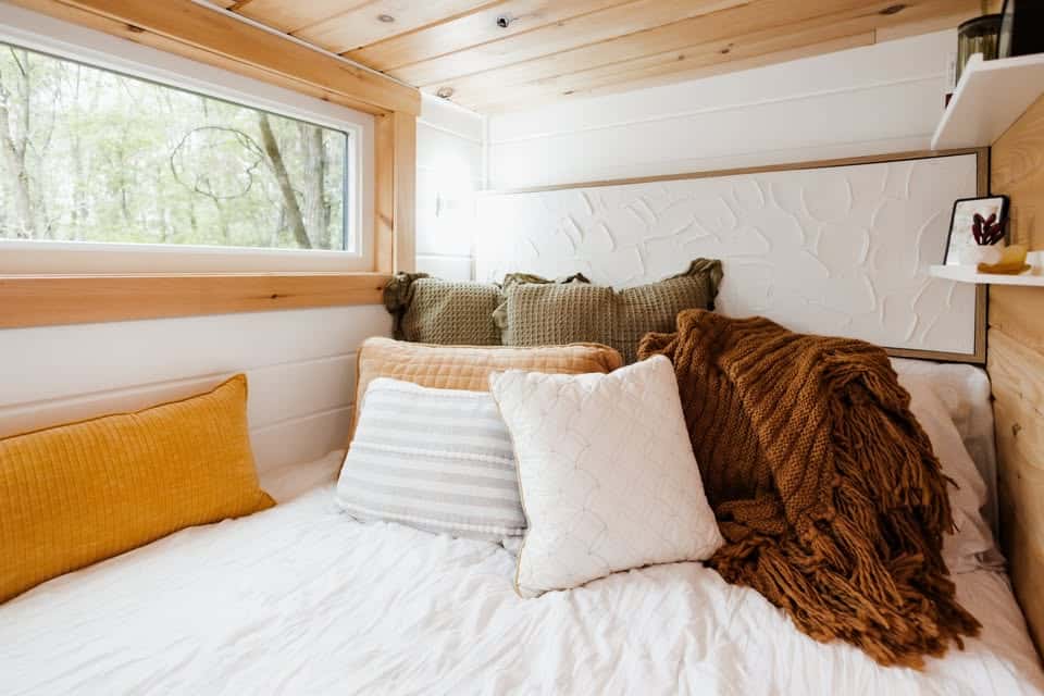 A cozy bed in a tiny house with white and striped pillows, a mustard yellow pillow, and a chunky brown throw blanket, set against a white textured headboard near a window with a view of trees.
