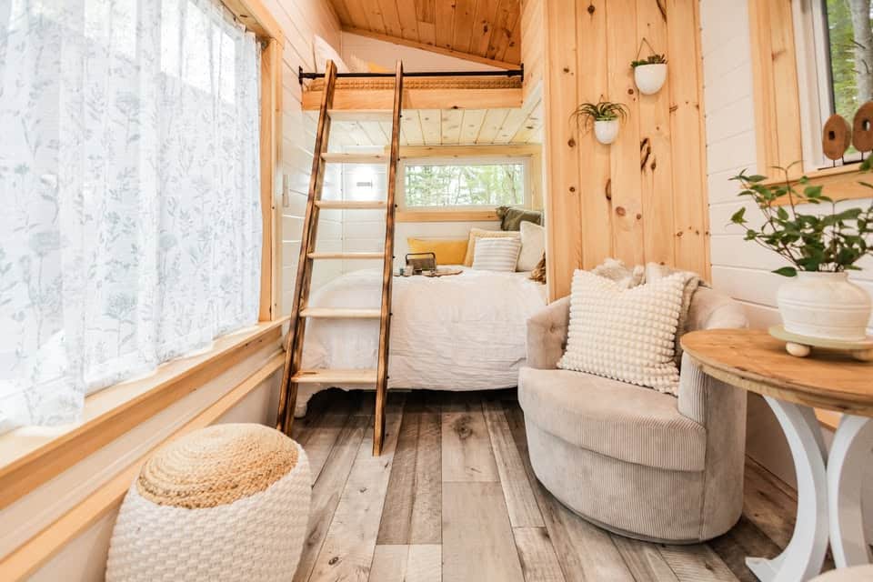 A cozy tiny house bedroom with wood-paneled walls, a bed with white bedding, a small loft and ladder, a round chair with pillows, a wooden side table with a plant, and sunlight streaming through floral curtains.