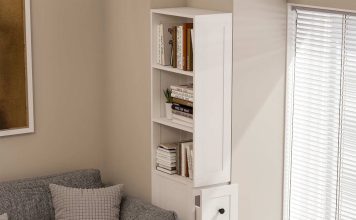 A narrow white bookcase with open shelves and three pull-out drawers stands beside a gray sofa and a window with blinds in a modern, light-filled living room.