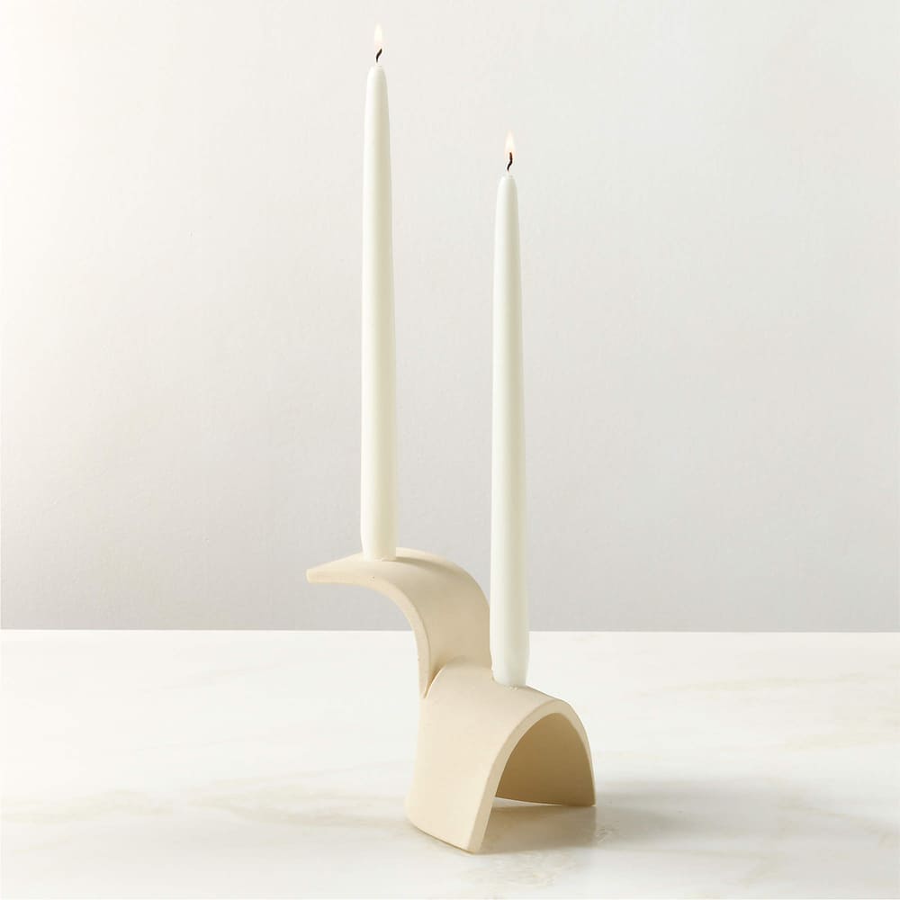 A modern, cream-colored ceramic candle holder with two white taper candles and a curved, sculptural design sits elegantly on a light marble surface against a plain white background.