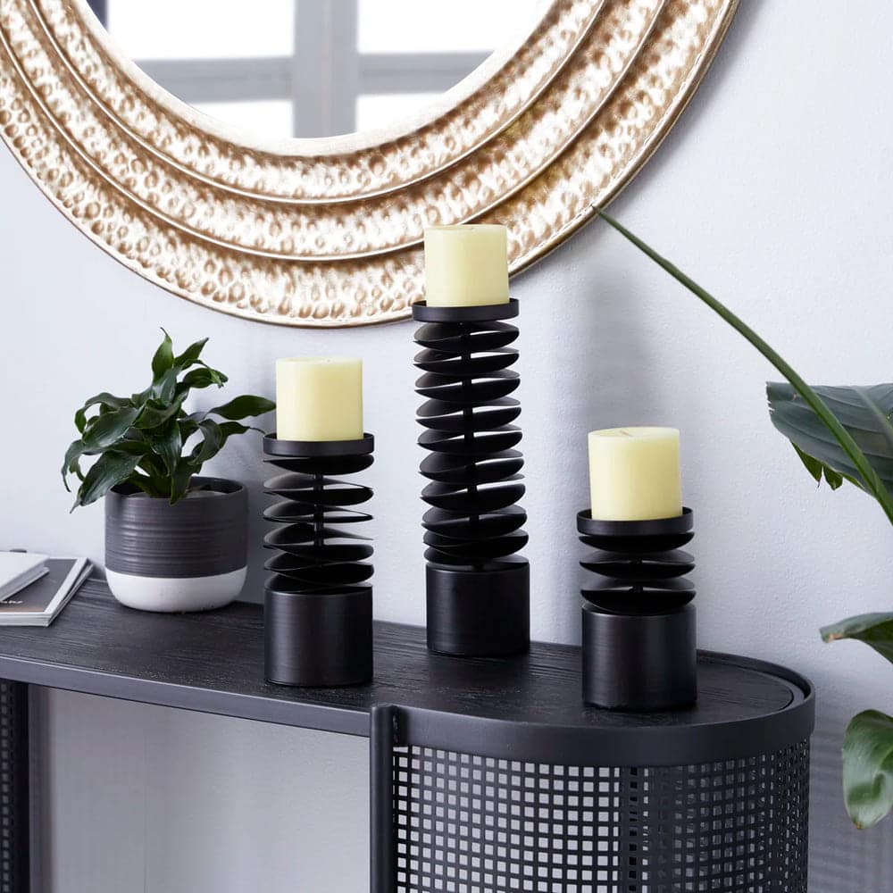 Three black spiral candle holders with light yellow candles are arranged on a black table. A potted plant and a large round gold-framed mirror add style to the space, highlighting the elegance of each candle holder.