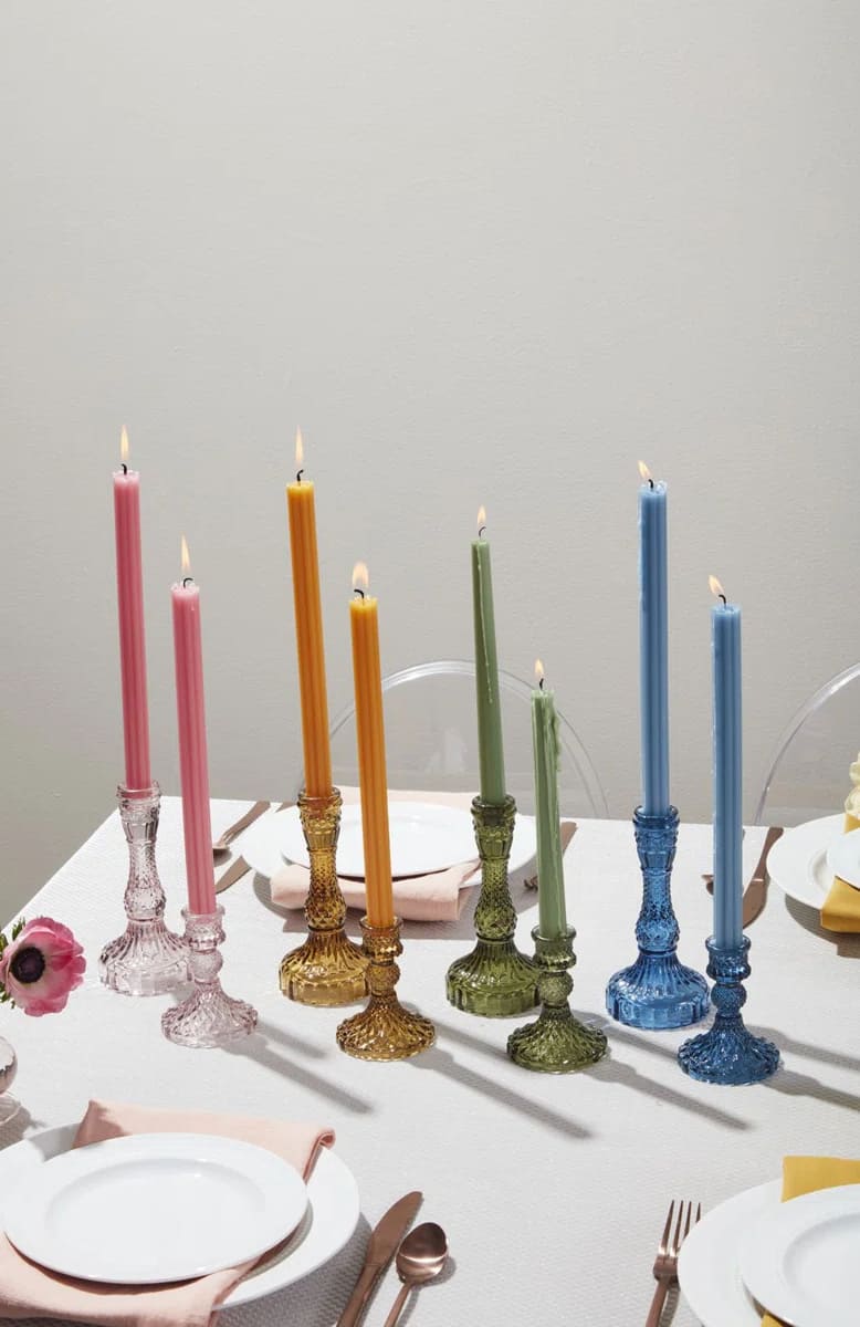 A set table with plates, cutlery, and napkins features six lit taper candles in pink, yellow, green, and blue, each glowing from an ornate candle holder that matches the color of the candle.