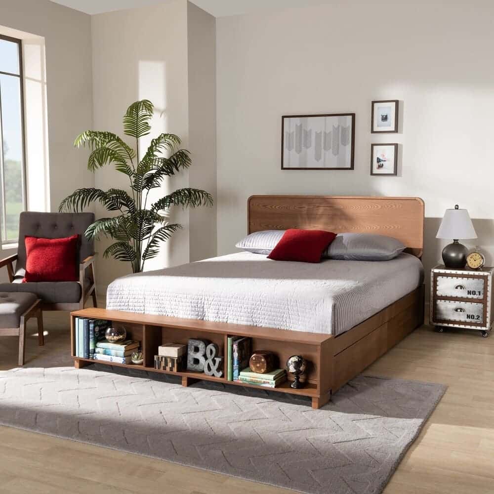 A modern bedroom features a storage bed with built-in shelves at the foot, white bedding, gray rug, armchair with red cushion, potted plant, nightstand, lamp, and framed wall art. Natural light fills the room.