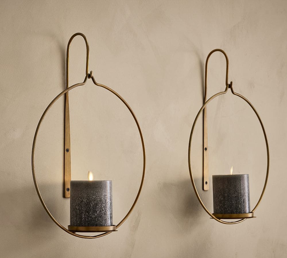 Two gold wall-mounted candle holders, each with a lit gray pillar candle, are displayed on a beige wall. The holders feature a modern, minimalist design that adds elegance to any space.