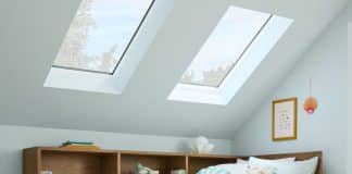 A cozy bedroom with a wooden storage bed featuring built-in shelves, pastel bedding, and pillows. Skylights let in natural light, and the walls are light blue. Books, baskets, and decorative items fill the shelves.