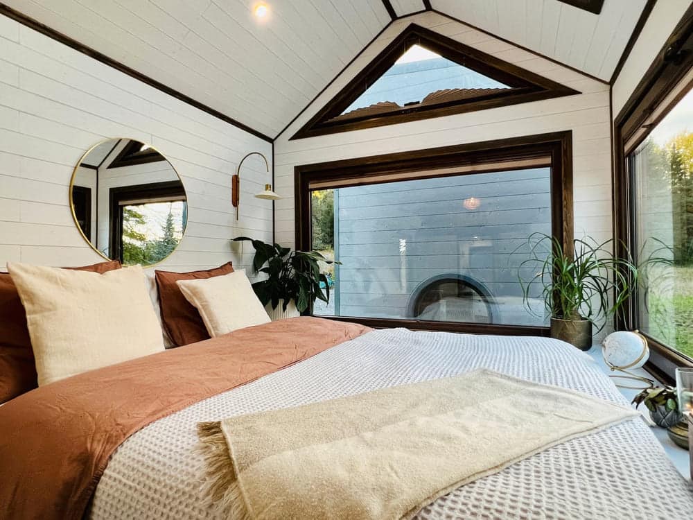 A cozy tiny house bedroom with white paneled walls, large windows, a bed with brown and white bedding, a round mirror, plants, and natural light streaming in.