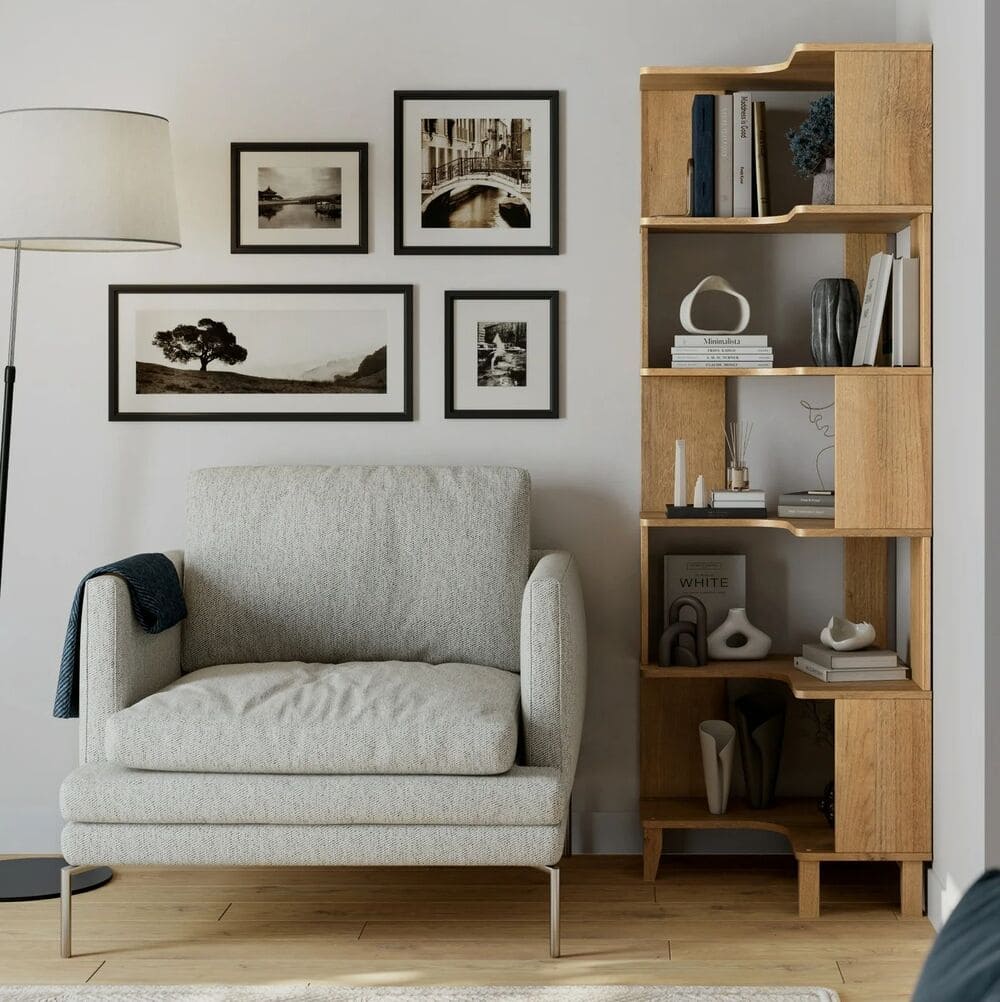 A cozy living room corner with a light gray armchair, a wooden bookcase filled with books and decor, a floor lamp, and a wall decorated with five framed black-and-white photos.