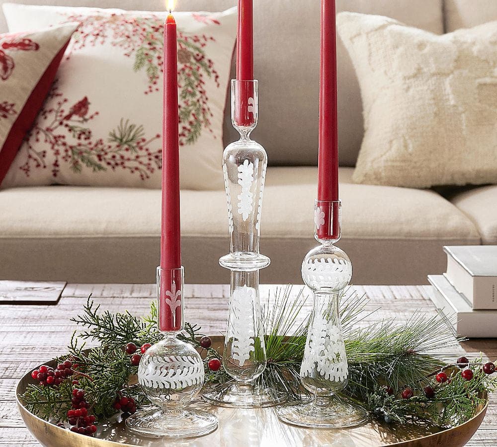 A decorative tray with three candle holders, each cradling a red candle, is surrounded by pine branches and red berries on a wooden table in front of a beige sofa with festive, nature-themed pillows.
