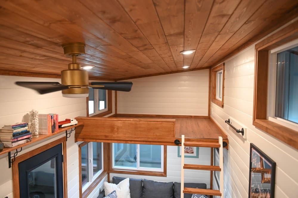 This cozy tiny house interior features wood-paneled ceilings, white walls, a loft area with a ladder, a ceiling fan, windows, a couch, and shelves with books and decor.