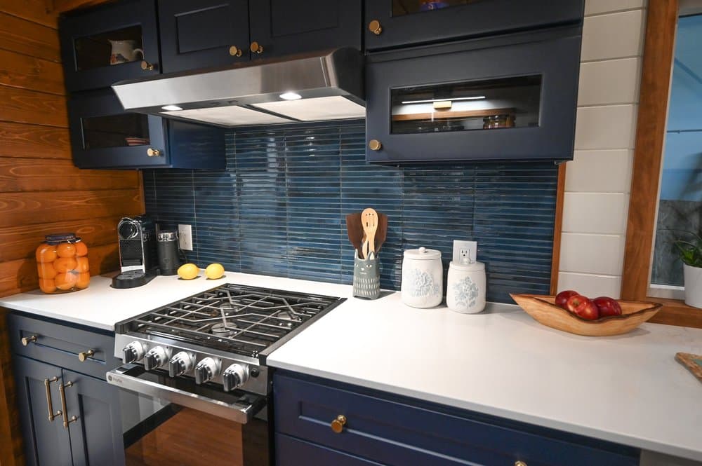 Modern tiny house kitchen with navy cabinets, a stainless steel gas stove, blue tile backsplash, white countertop, coffee maker, jars, wooden utensils, lemons, a bowl of apples, and a bowl of oranges on the counter.