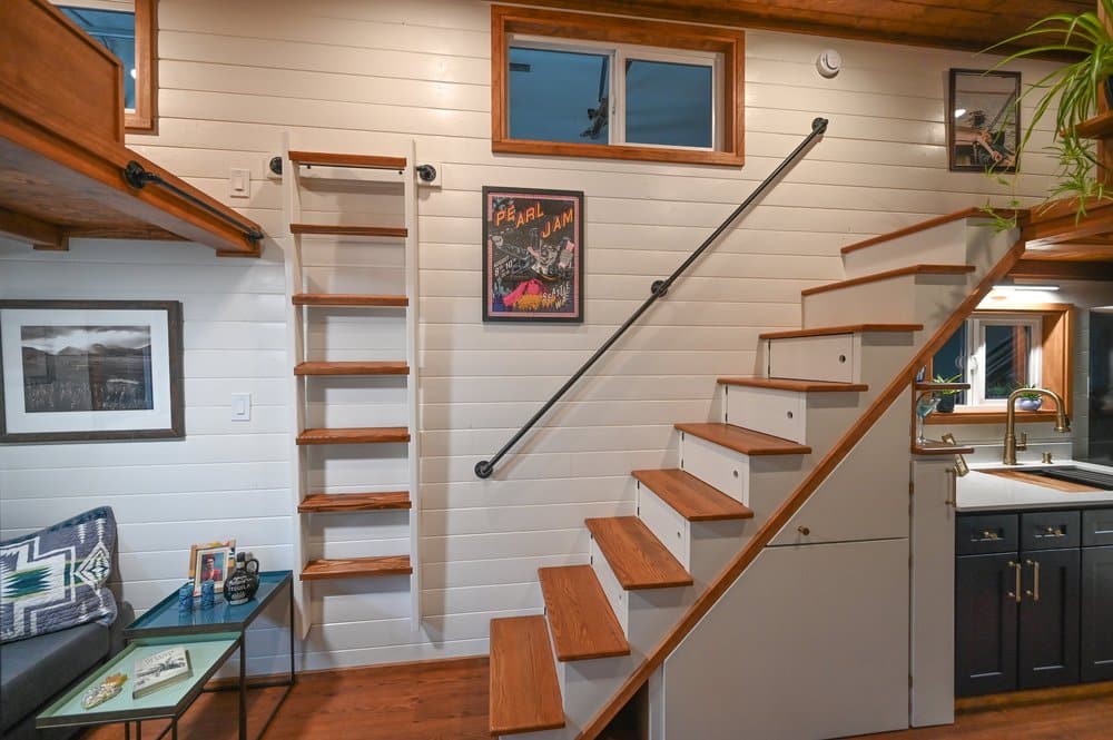 A cozy tiny house interior with white shiplap walls, wooden stairs with black railing, a ladder to a loft, framed art and photographs, a small sitting area, and a dark kitchen counter with a sink.