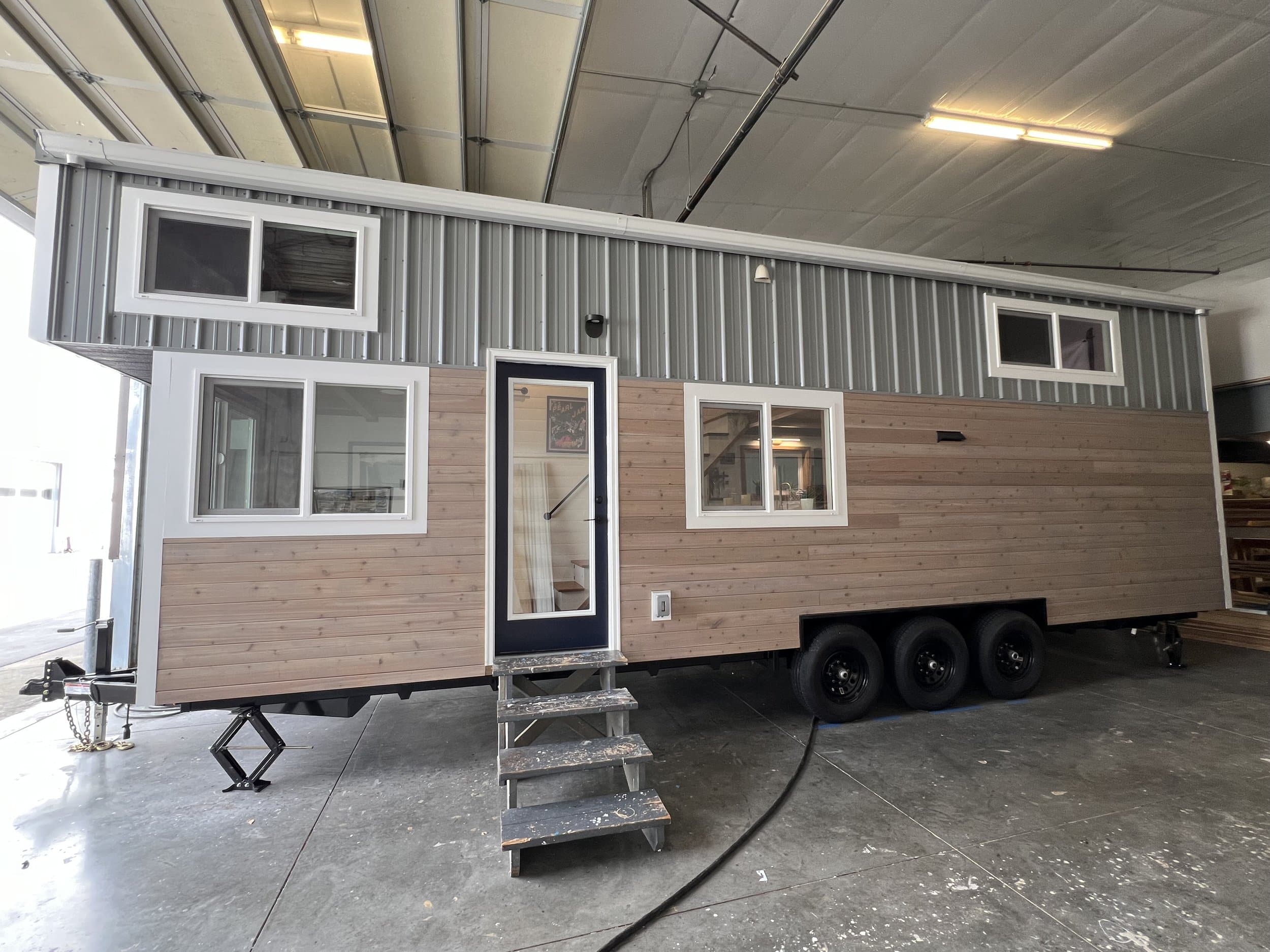 A modern tiny house on wheels with wood siding, large windows, and a black door, parked inside a spacious, well-lit garage or workshop—perfect for those seeking the freedom of tiny house living.
