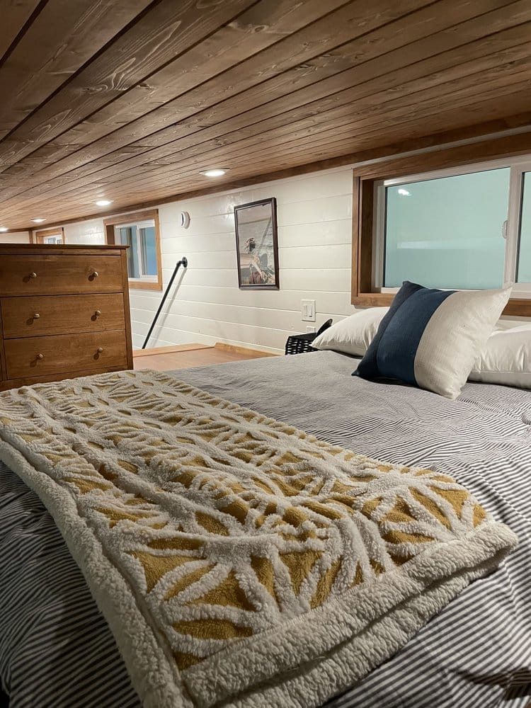 A cozy tiny house bedroom with a low wooden ceiling, striped bedding, decorative pillows, a textured yellow and white blanket, a wooden dresser, wall art, and windows letting in natural light.