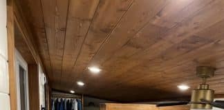 A compact, modern tiny house interior with wooden floors and ceiling, a kitchen below, and a cozy loft bedroom above accessed by wooden stairs. Plants and books decorate the space, and a window fills the tiny house with natural light.