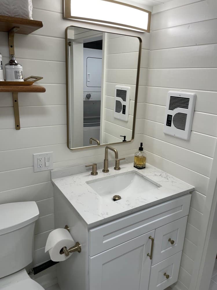 A modern white tiny house bathroom features a marble countertop, gold faucet, rectangular mirror, wall-mounted light, shelves, toilet, and wall heater. Soap dispenser and toilet paper are visible against crisp white shiplap walls.