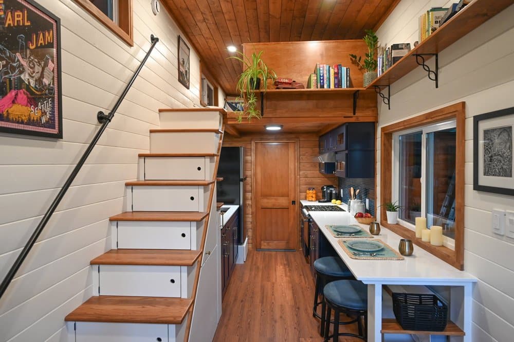A cozy tiny house interior with wooden floors and ceiling, a staircase with storage drawers, a kitchen on the right, bar seating with place settings, wall art, shelves filled with plants and books, and a door at the far end.