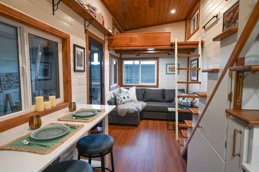 A cozy, modern tiny house interior with white walls, wood ceiling, a loft bed accessible by ladder, gray sectional sofa, and a small dining area set for two with green placemats.