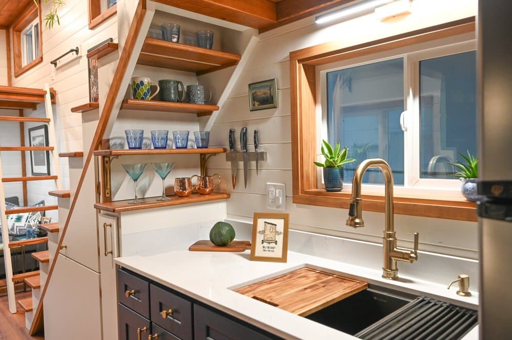 Modern tiny house kitchen with open wood shelves, glassware, a knife rack, gold faucet, plants, a window, and stairs leading up; wooden accents and white shiplap walls create a cozy, organized tiny house space.