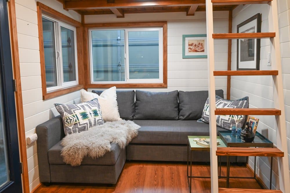 Cozy tiny house living room with a gray sectional sofa, decorative pillows, and a furry throw. A small glass side table holds magazines beneath a wooden loft ladder. White walls and wooden trim frame large windows.
