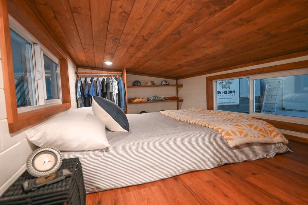 A cozy tiny house bedroom with a low wooden ceiling, striped bedding and pillows on the bed, an alarm clock on a nightstand, and a small open closet. Large windows fill the space with natural light.