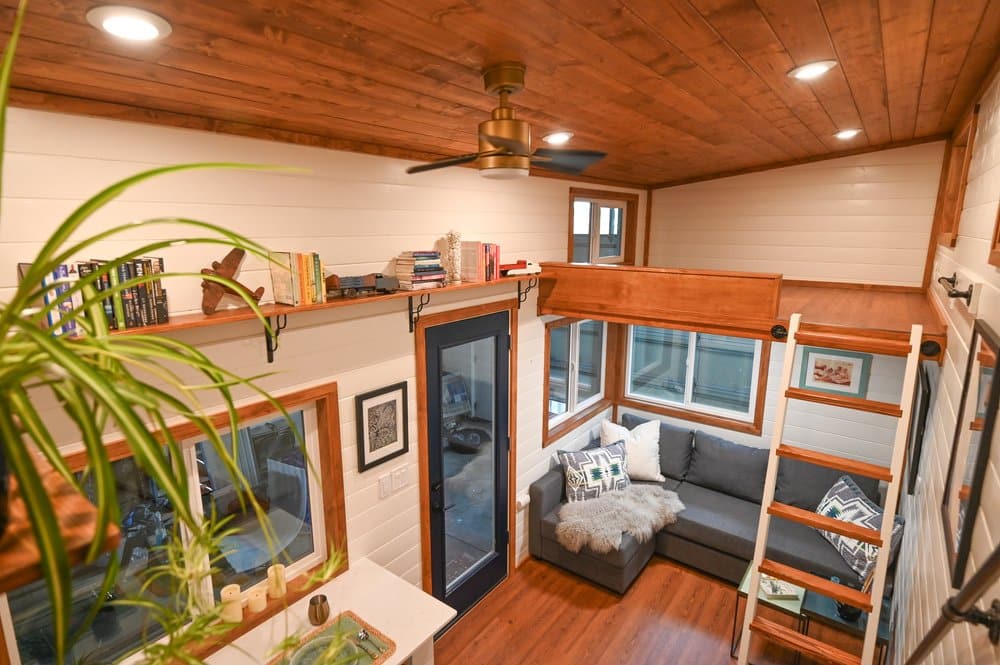 A cozy tiny house interior with wood floors and ceiling, a loft accessible by ladder, a gray sectional sofa by large windows, shelves with books, and a ceiling fan above.