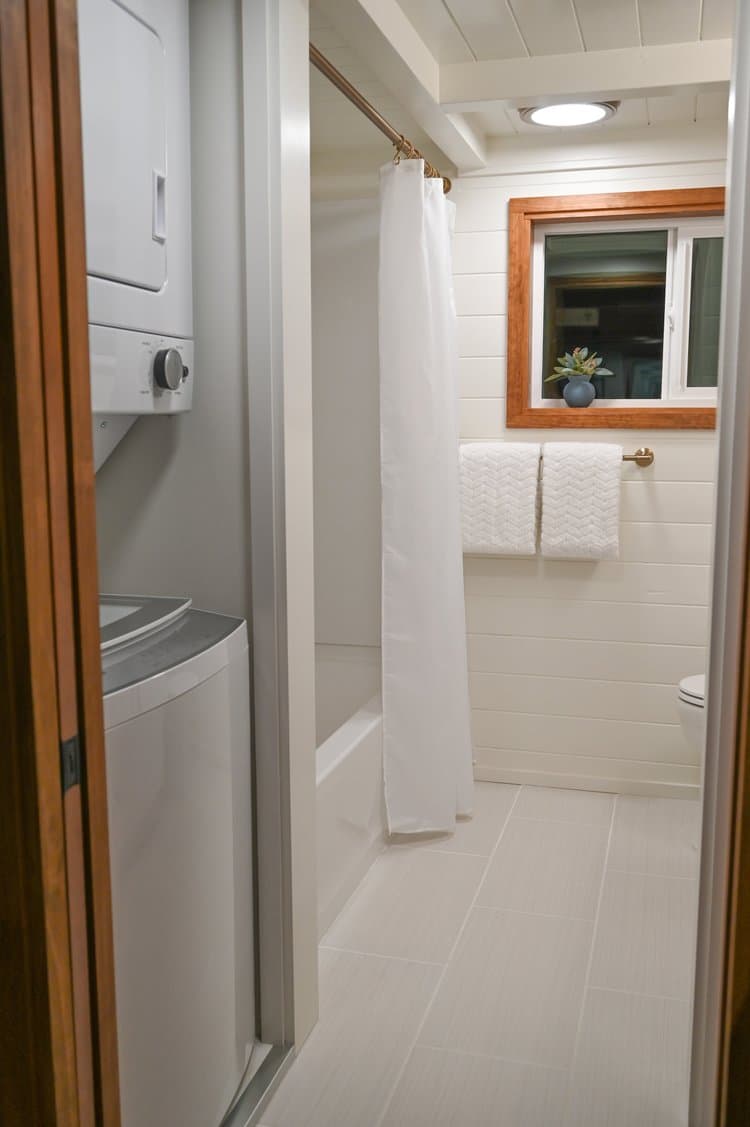 Small bathroom in a tiny house with a stacked washer and dryer, white shower curtain over a bathtub, two white towels on a gold towel rack, a window with a wood frame, and a small plant on the windowsill.