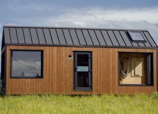 A modern tiny house with a dark metal roof and large windows sits on a grassy field, surrounded by hills and trees under a partly cloudy sky.