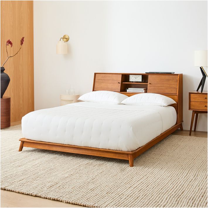 A modern bedroom with a stylish storage bed featuring built-in shelves in the headboard, white bedding, a beige textured rug, wooden nightstand, lamp, and minimalist decor in neutral tones.