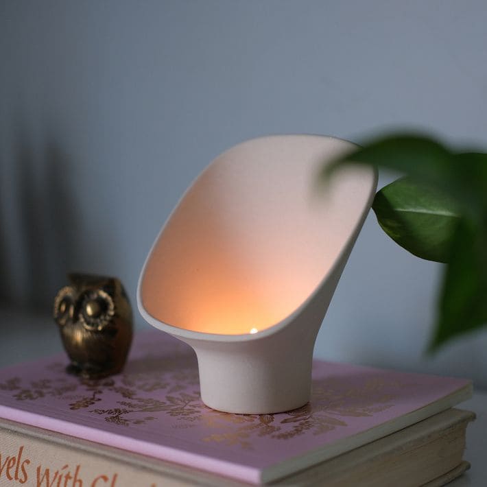 A small, modern white lamp with a soft orange glow sits atop a pink and gold book, next to a candle holder shaped like an owl figurine and a green plant in the foreground.