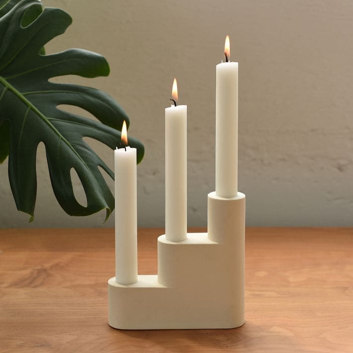 Three white candles of varying heights are aglow in a modern candle holder on a wooden surface. A large green monstera leaf peeks out in the background.