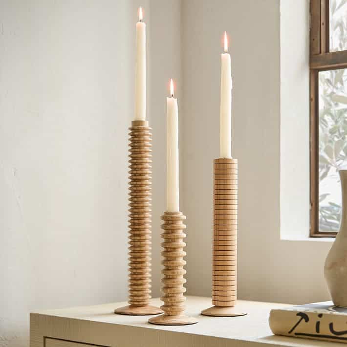 Three lit candles in tall, ribbed candle holders are arranged on a light-colored surface near a window. The scene has a minimal, modern aesthetic with soft natural lighting.