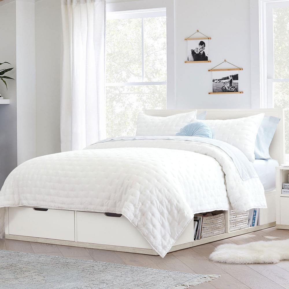 A bright bedroom with large windows, a white storage bed featuring a quilted comforter, pale blue accent pillow, and built-in shelves beneath. Minimalist decor includes wall art and a soft rug on a light wood floor.