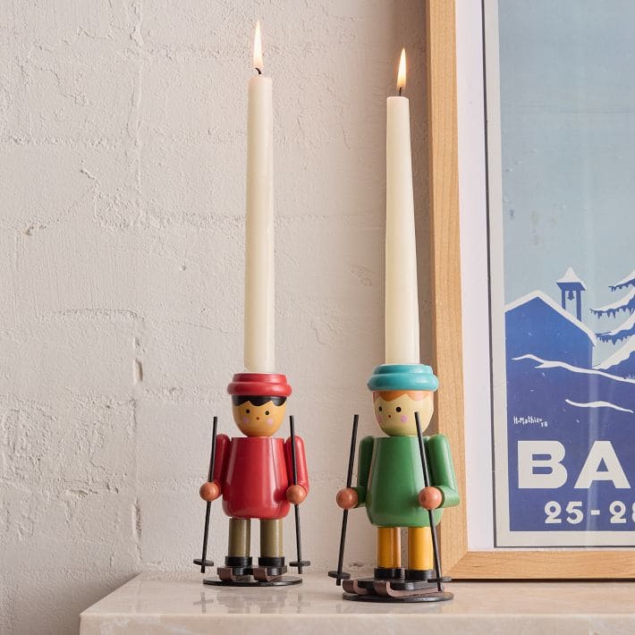 Two candle holders shaped like wooden skiers, one in red and one in green, each holding ski poles and topped with lit white candles. They sit on a mantel beside a framed winter poster.