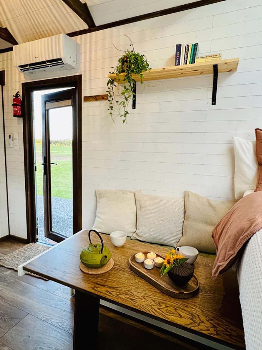 A cozy nook in a tiny house with a wooden table set for tea, featuring a green teapot, cups, and a tray with candles and flowers. A couch with cream cushions sits below books and a hanging plant, as natural light streams through the open door.