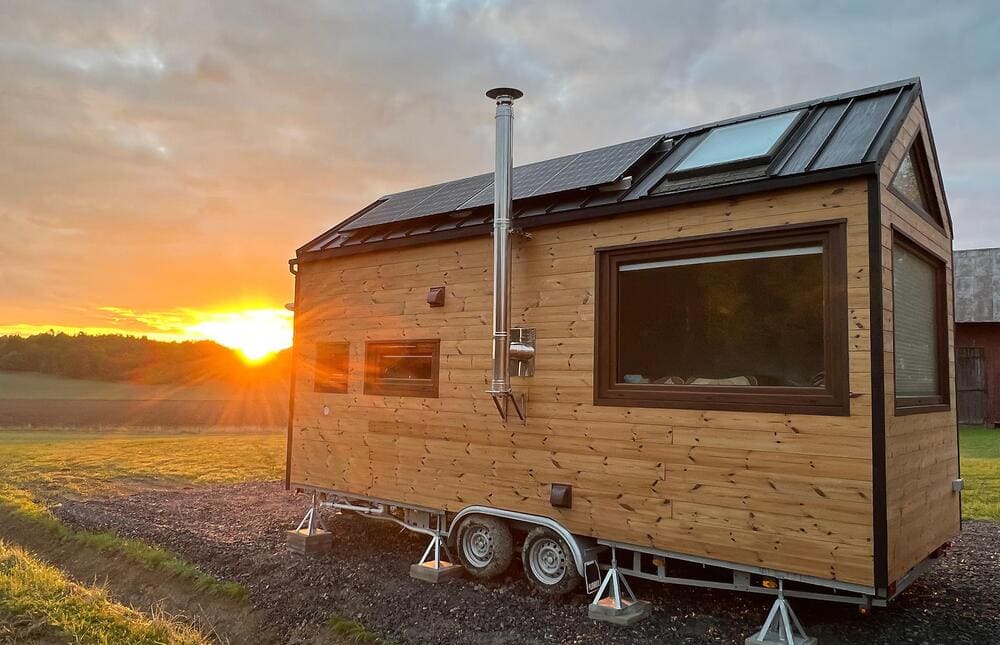A small wooden tiny house on wheels with large windows and a chimney sits on gravel in a field at sunset, its solar panels gleaming atop the roof beneath a vibrant sky.