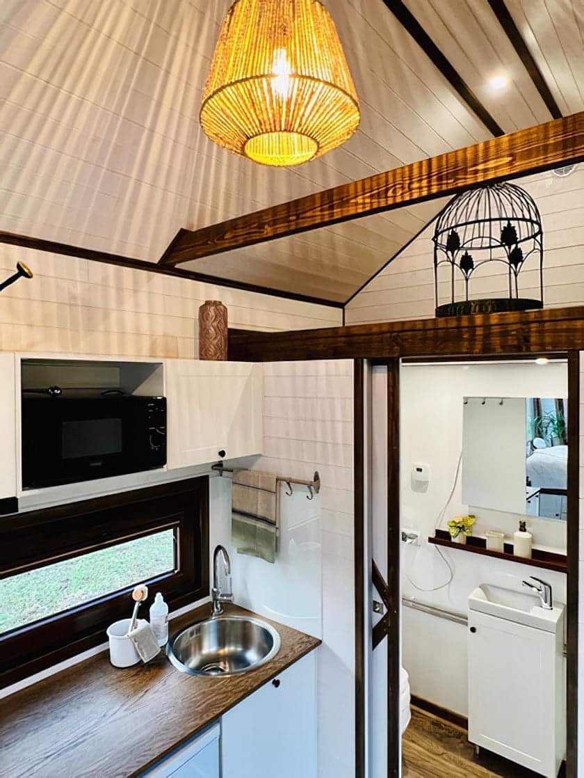 A cozy, modern tiny house kitchen with a sink, microwave, and towel rail. Above, a rattan pendant light hangs from the vaulted wooden ceiling. A doorway leads to a compact tiny house bathroom with a washing machine and mirror.