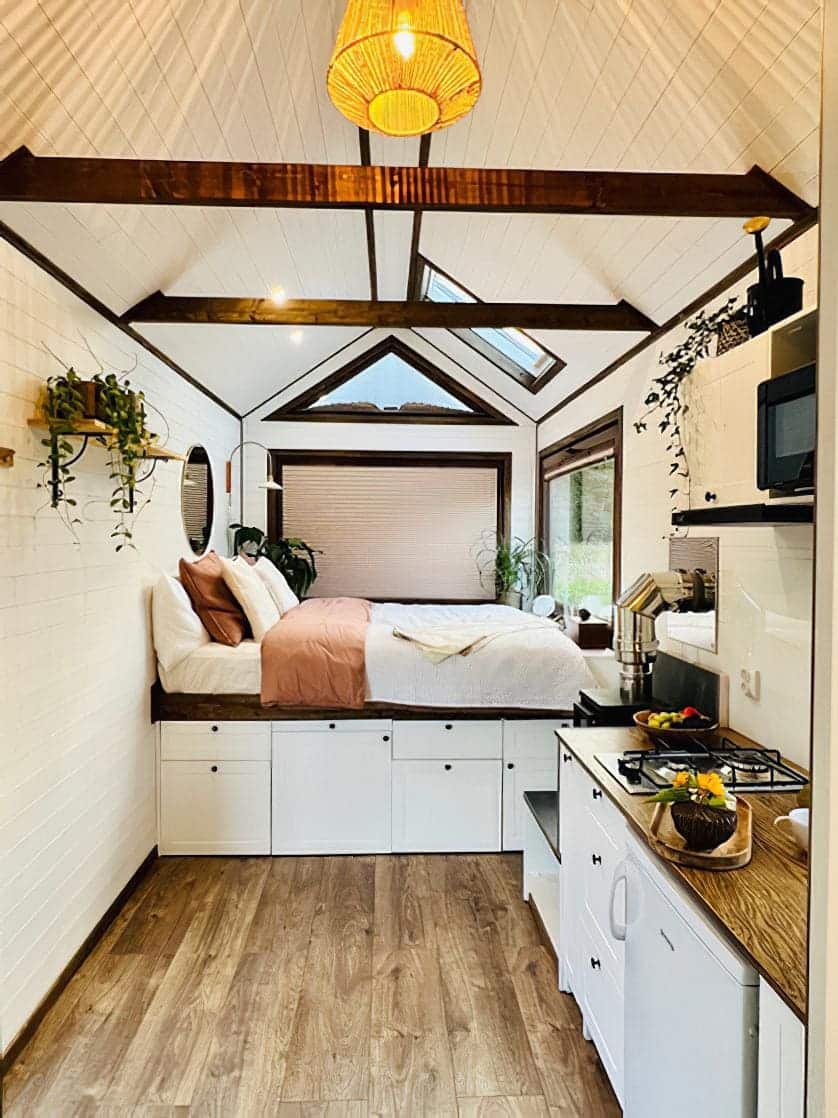 A cozy tiny house interior with a raised bed, white cabinets, wood accents, potted plants, a small kitchen area, and large windows letting in natural light. Warm lighting and neutral decor create an inviting tiny house atmosphere.